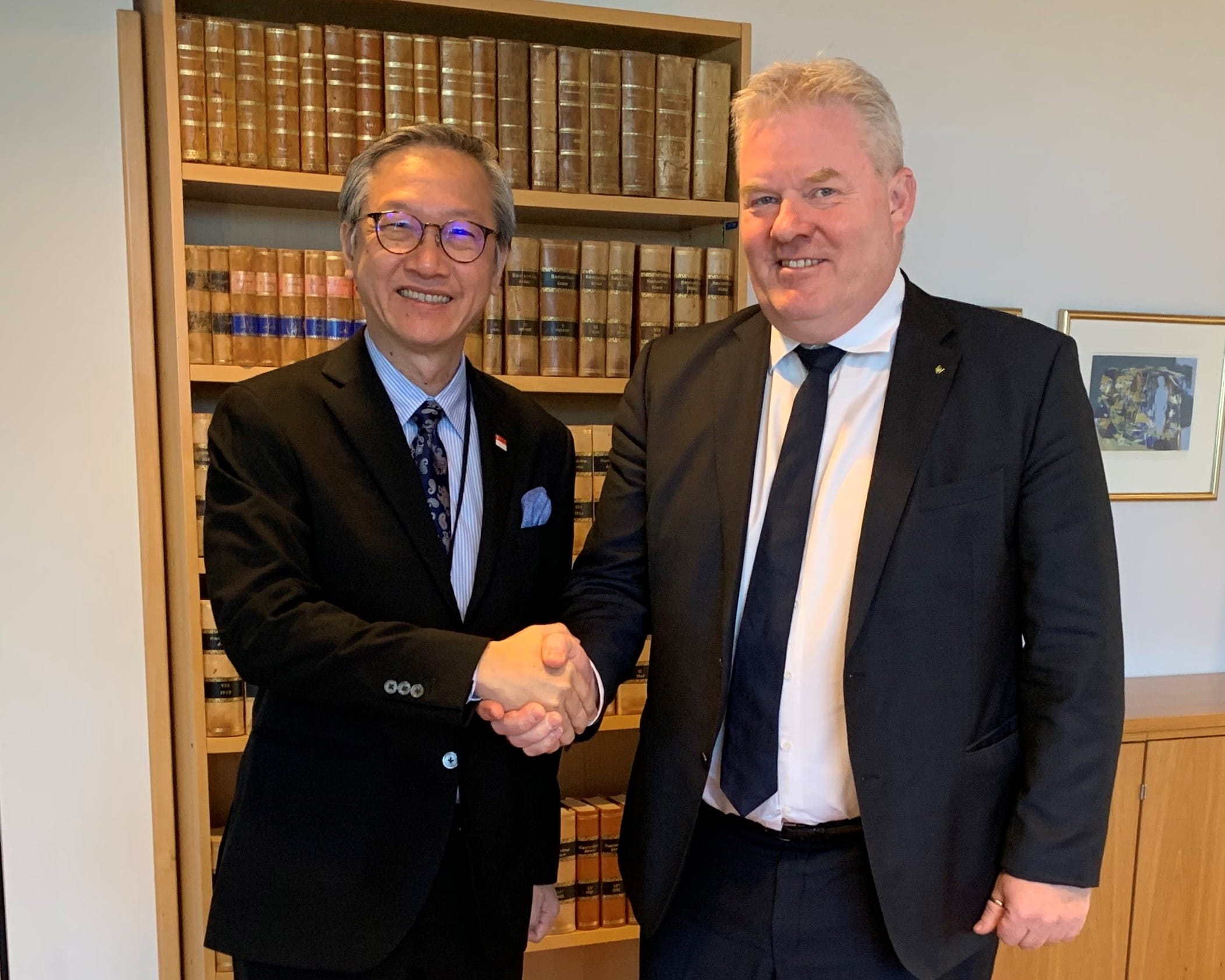 Two men in suits shaking hands in front of a bookcase filled with books.